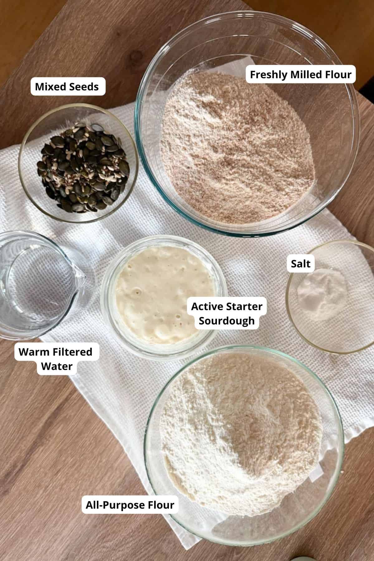 Image shows fresh milled flour, mixed seeds, active sourdough starter, all-purpose flour, salt, warm filtered water on a wooden surface.