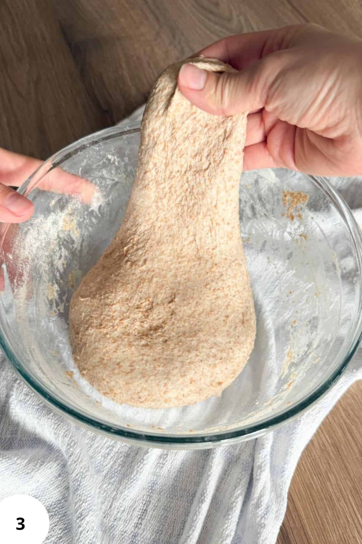 Hand holding a piece of freshly milled flour bread dough over a glass bowl.