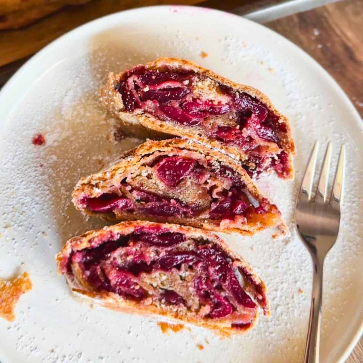 Close-up of cherry strudel slices on a white plate with a fork.