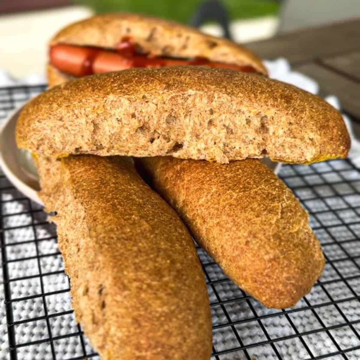 Freshly baked sourdough hot dog buns on a cooling rack with a hot dog in the background.