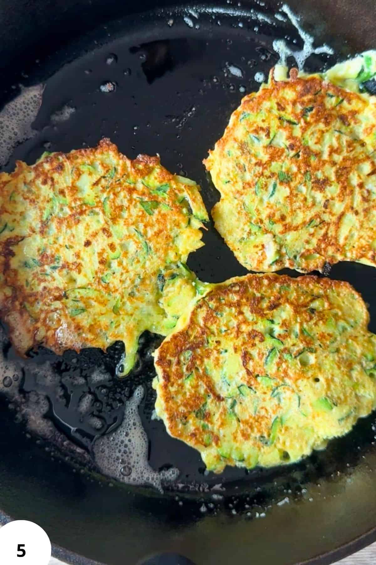 Golden zucchini fritters cooking in a skillet with green herbs.